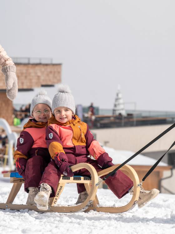 Zwei Kinder sitzen lachend auf dem Schlitten im Winterurlaub in den Alpen