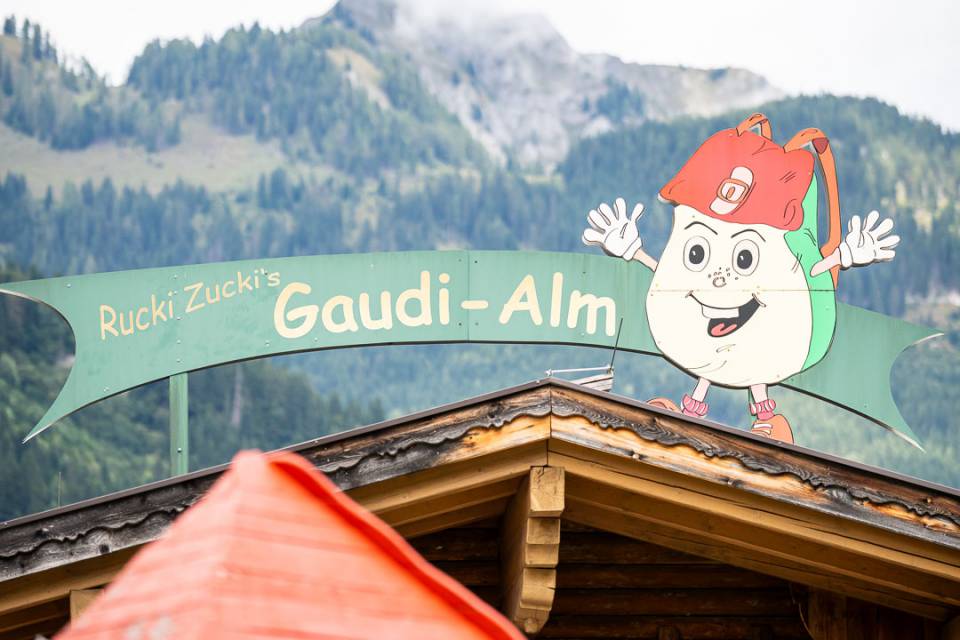 Holzhütte mit Schild „Rucki Zucki’s Gaudi-Alm“ und Bergkulisse im Hintergrund