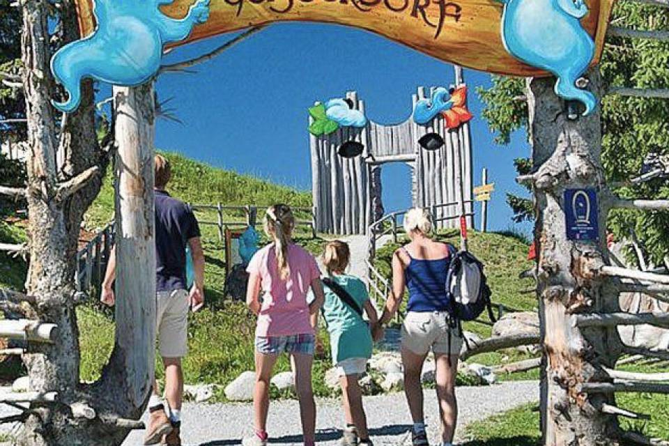 Familie geht durch ein Holztor mit der Aufschrift „Willkommen im Geisterdorf“ auf einem Erlebnisweg