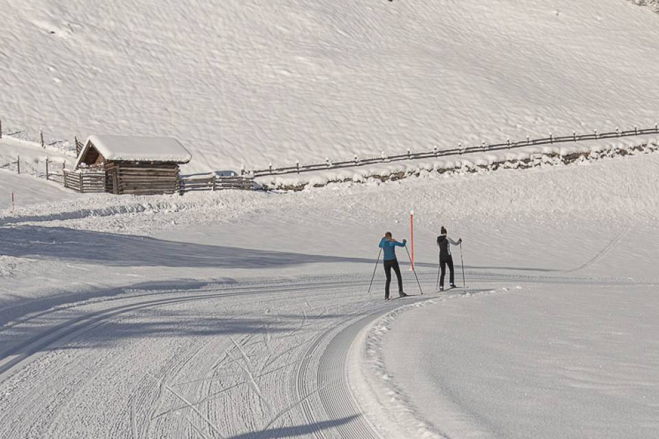 Zwei Personen fahren auf einer verschneiten Loipe durch eine winterliche Landschaft mit Holzhütte – Langlaufen rund um das Familienhotel Oberkarteis