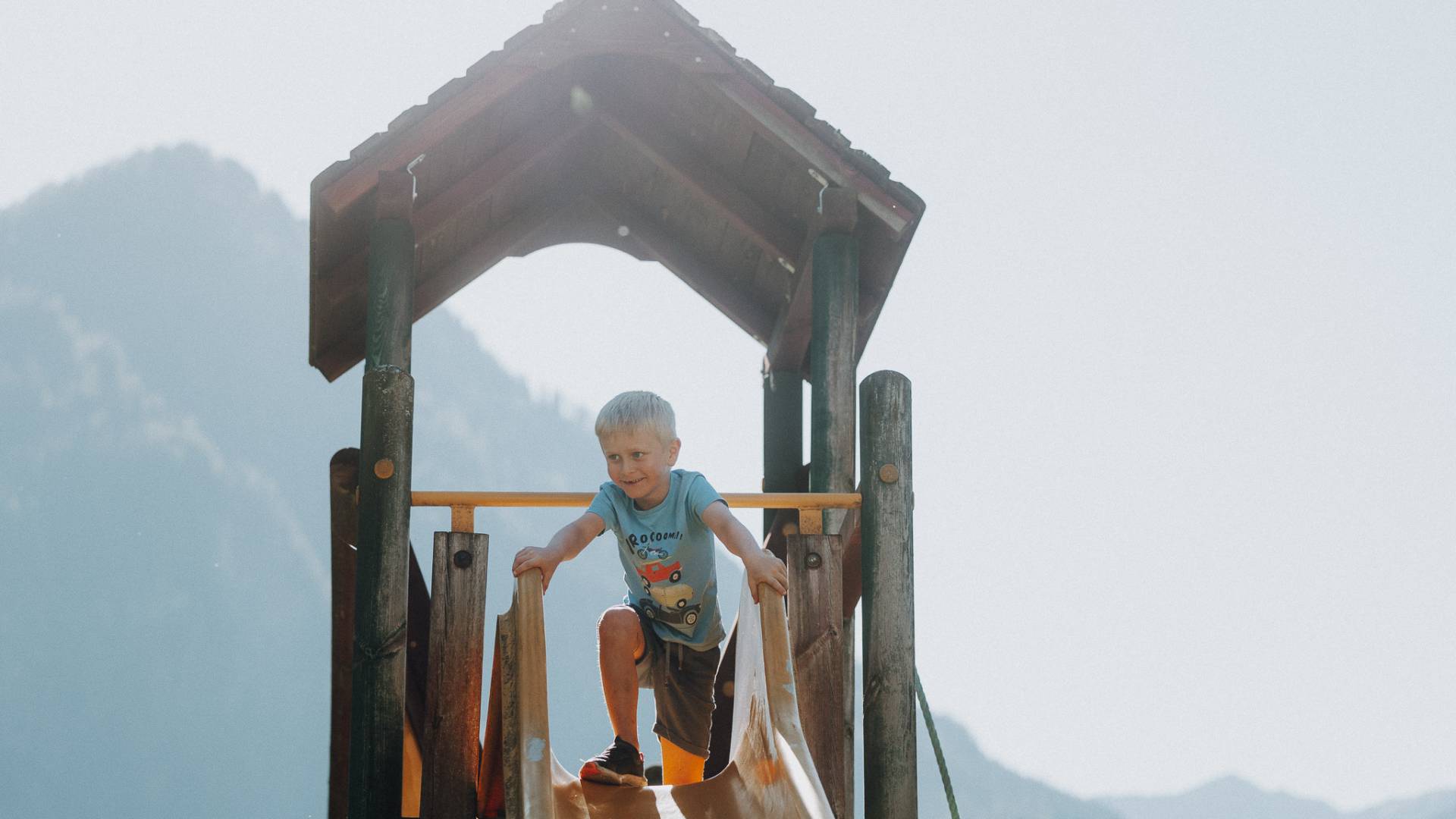 Junge steht oben auf der Rutsche des Spielturms mit Bergpanorama im Hintergrund im Familienhotel Oberkarteis