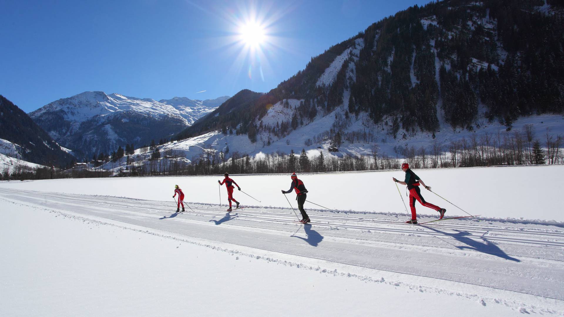 Vier Langläufer gleiten bei Sonnenschein über eine präparierte Loipe vor verschneiten Bergen – Wintererlebnis nahe dem Familienhotel Oberkarteis
