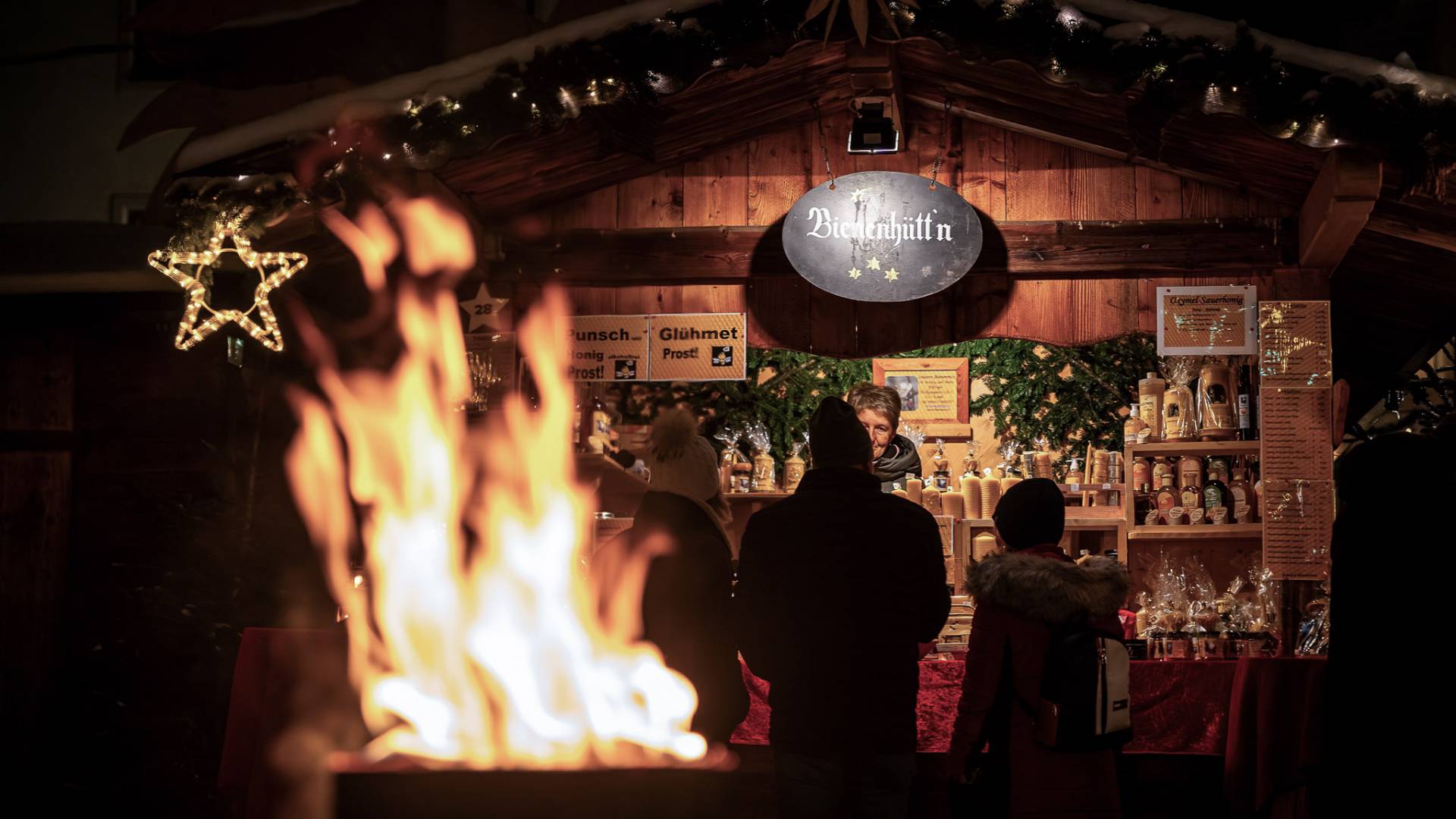 Weihnachtsmarkthütte mit Lichterketten und Feuerstelle im Vordergrund, Besucher stehen vor dem Stand – Winterstimmung rund um das Familienhotel Oberkarteis