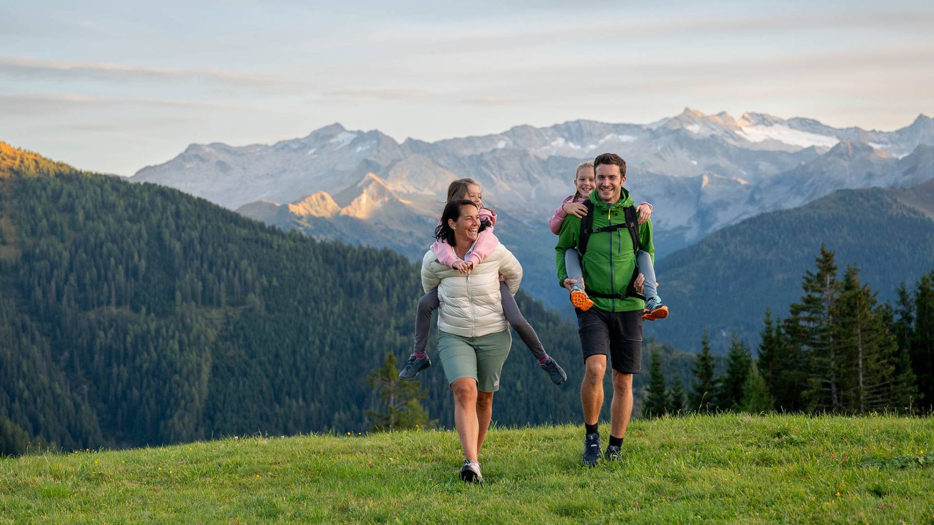 Familie mit Kindern huckepack auf Almwiese mit Alpenpanorama im Großarltal